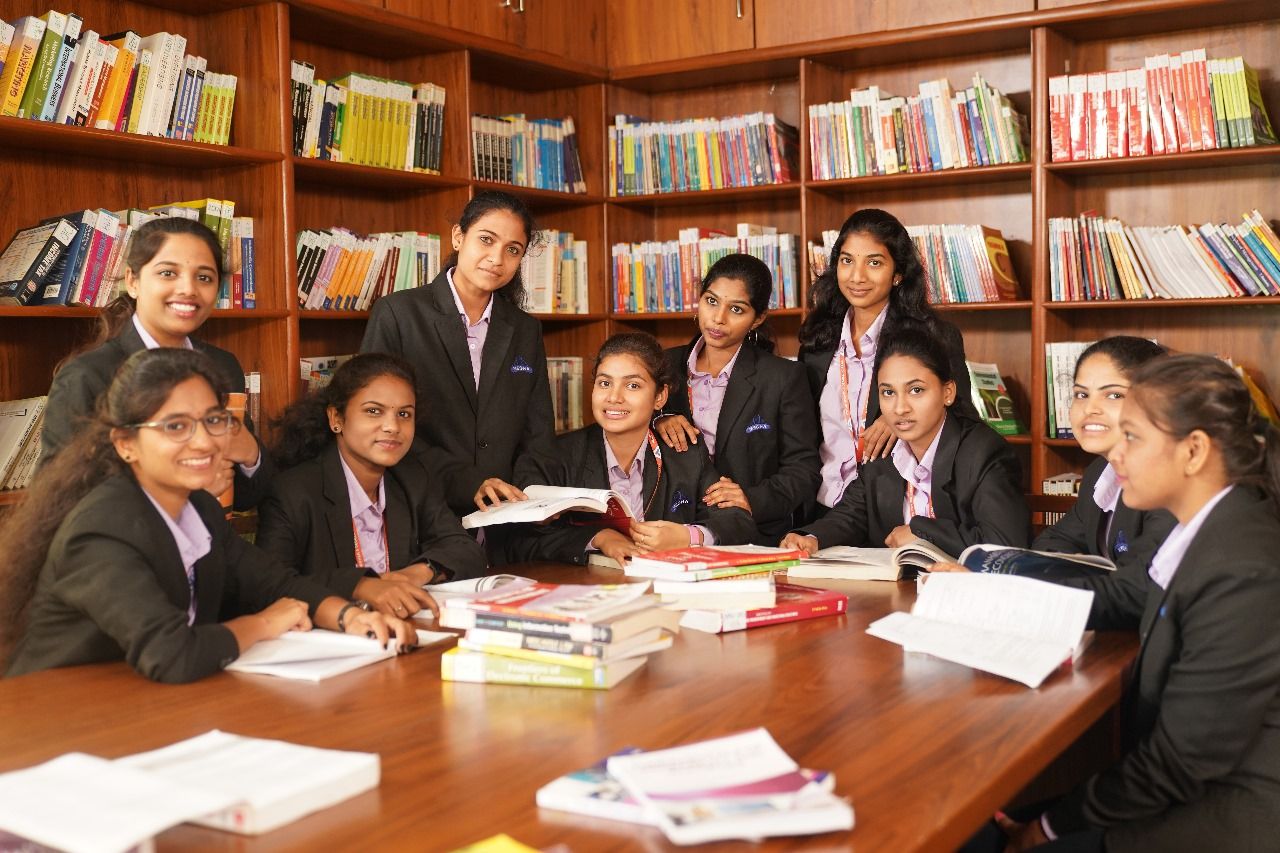 Students in blazers around a library reading table with open books, bookshelves behind them